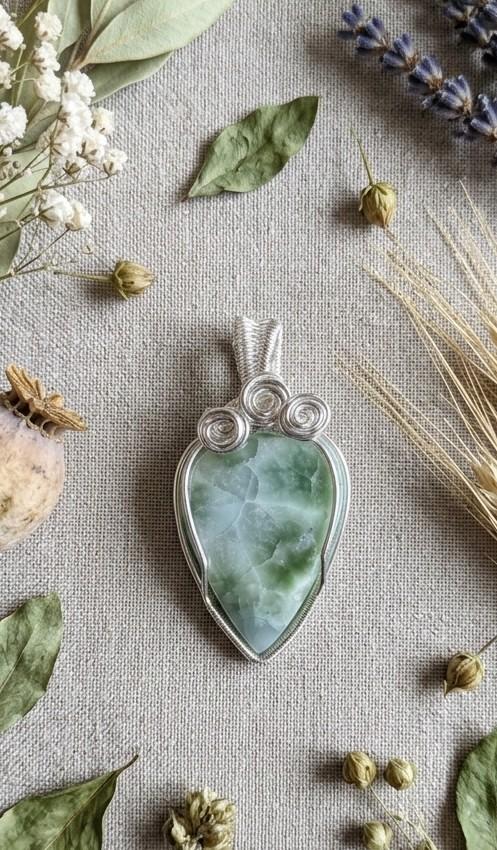 Handgemaakte verzilverde wire-wrap hanger met een druppelvormige groen-blauwe hemimorfiet steen, gedecoreerd met drie zilveren krullen, gepresenteerd op een linnen ondergrond.