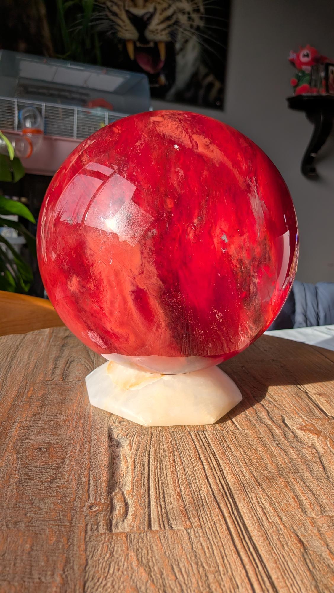 Een gigantische, glanzend rode smeltkwarts bol van 22 cm diameter en 12.05 kilo met dynamische interne patronen, gepresenteerd op een witte zeshoekige houder op een houten tafel.