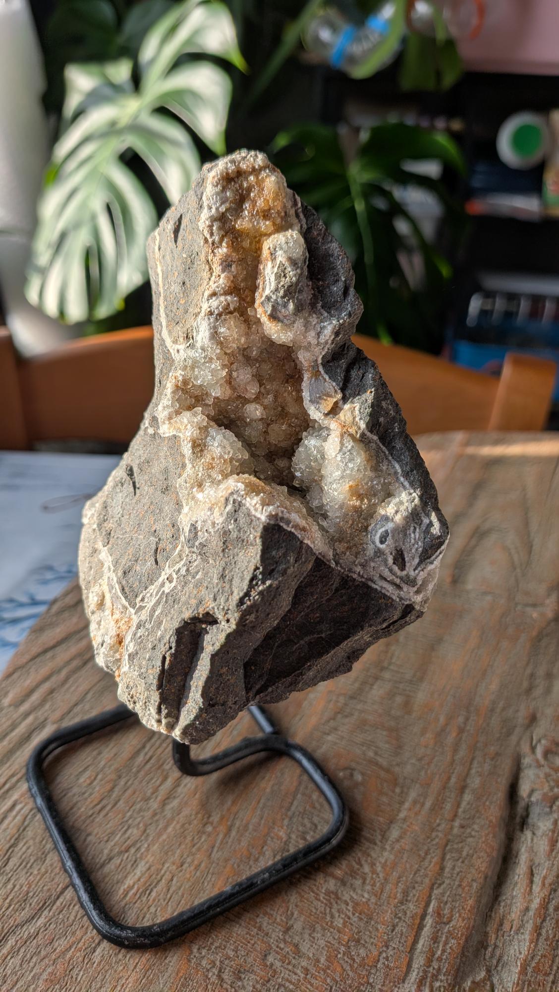 Een ruwe, donkere gesteente-geode met een opening vol witte calcietkristallen, gepresenteerd op een minimalistisch zwart metalen statief.