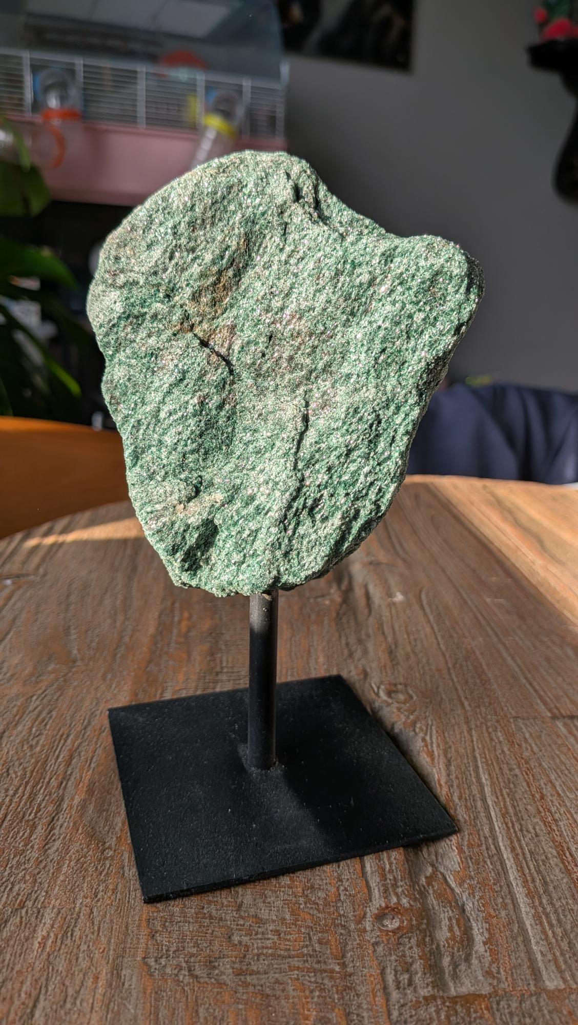 Een ruw, diepgroen stuk fuchsiet met een zilverachtige glinstering, gepresenteerd op een modern zwart metalen statief tegen een lichte achtergrond.