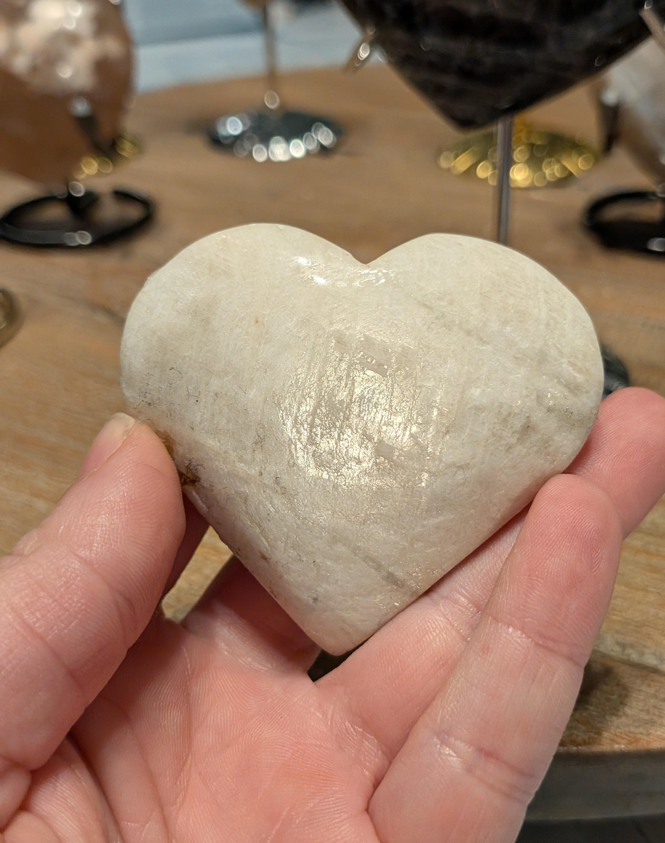 "Handgepolijst perzik-wit maansteen hart van 115 gram vastgehouden in een hand, toont de afmeting van 6.5 cm."