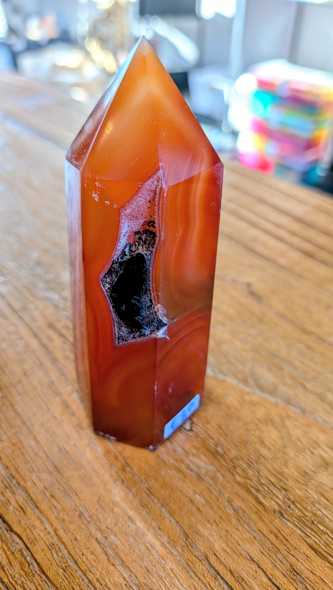 "Gepolijste oranje carneool toren van 10 cm hoog met een opvallende, donkere natuurlijke kristalholte in het midden op een houten ondergrond."