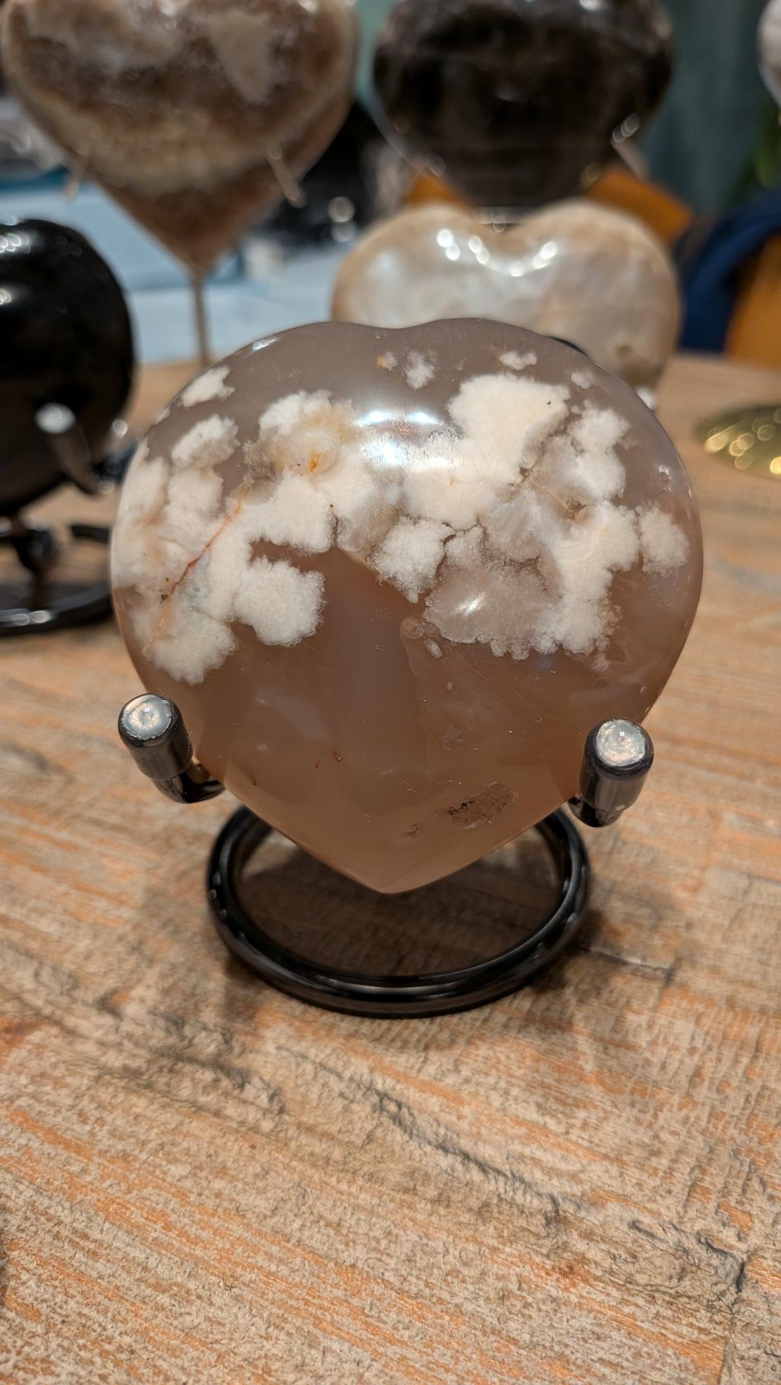 "Handgeslepen Bloemagaat hart van 182 gram op een zwarte standaard, tonend de witte bloem-insluitingen."