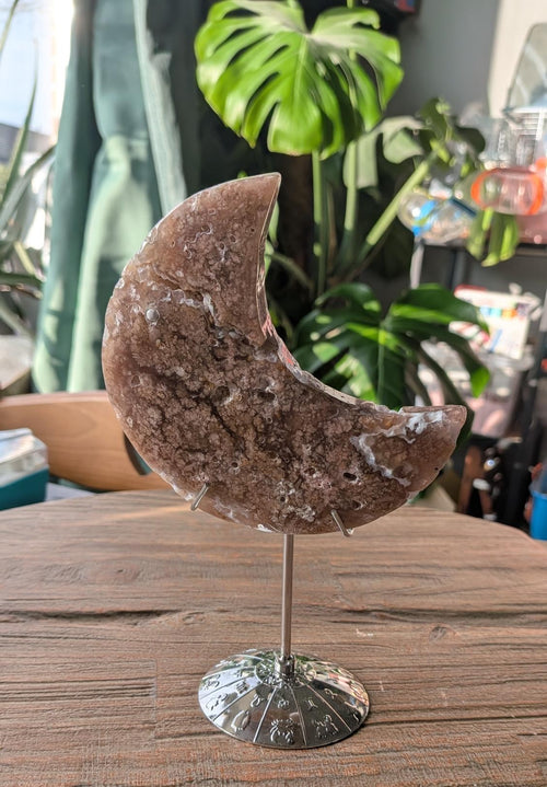 Handgeslepen halve maan van Bloesem Agaat (268 gram) gepresenteerd op een zilverkleurige metalen standaard met gegraveerde sterrenbeelden, tonend een rijk patroon van roze en beige bloemachtige chalcedoon-insluitingen tegen een achtergrond met kamerplanten.