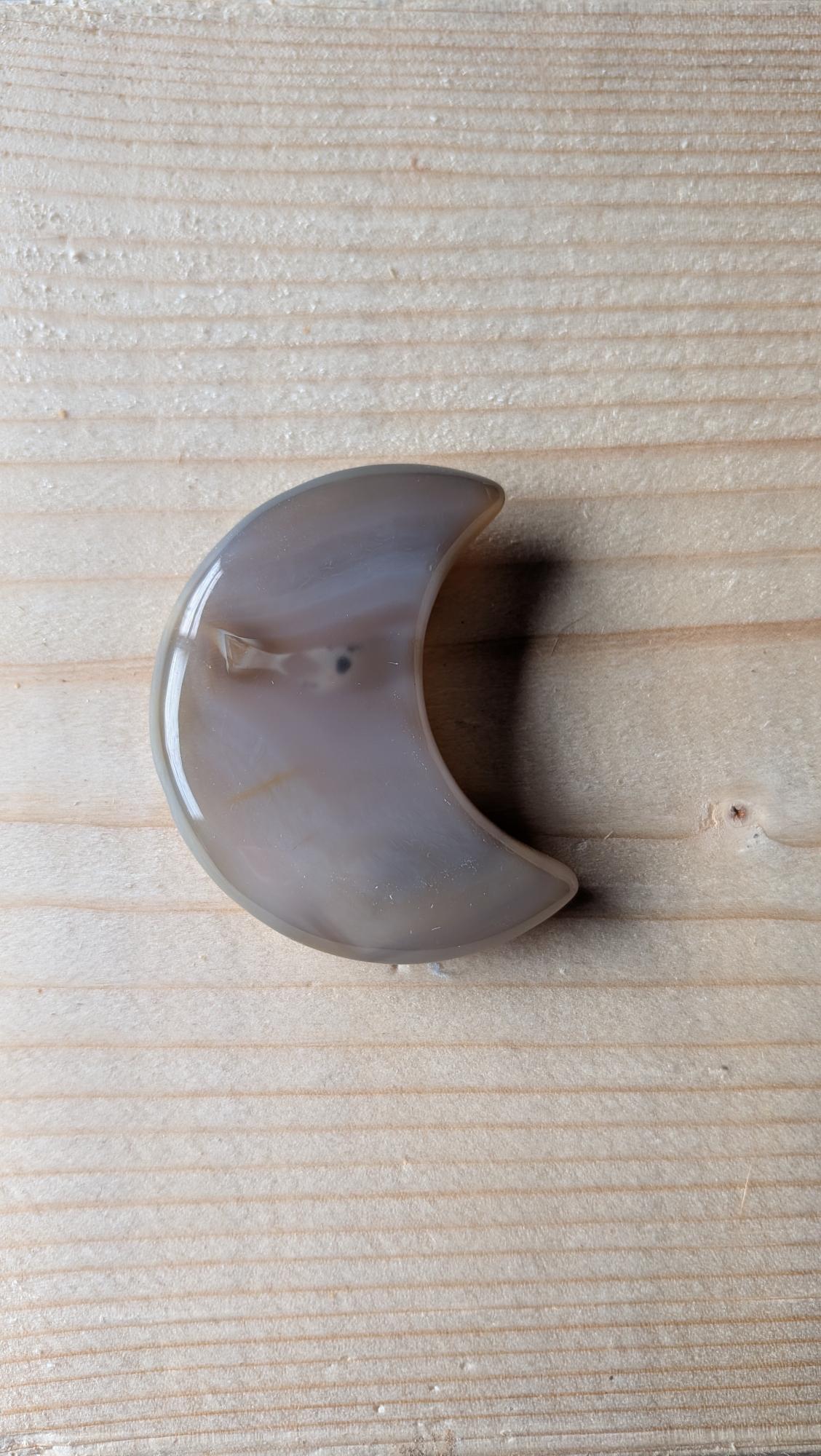 Handgeslepen Agaat edelsteen in de vorm van een halve maan, met zachte grijze en bruine kleurbanden, gefotografeerd op een houten achtergrond.