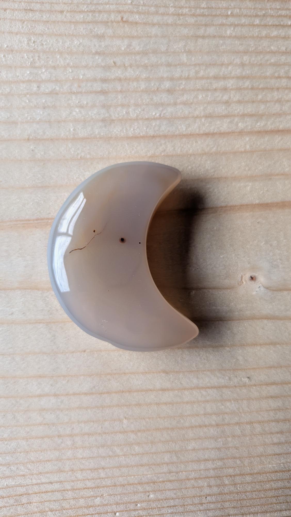 Een gepolijste grijze Agaat edelsteen in de vorm van een halve maan, 5 centimeter groot, gefotografeerd op een lichte houten ondergrond.