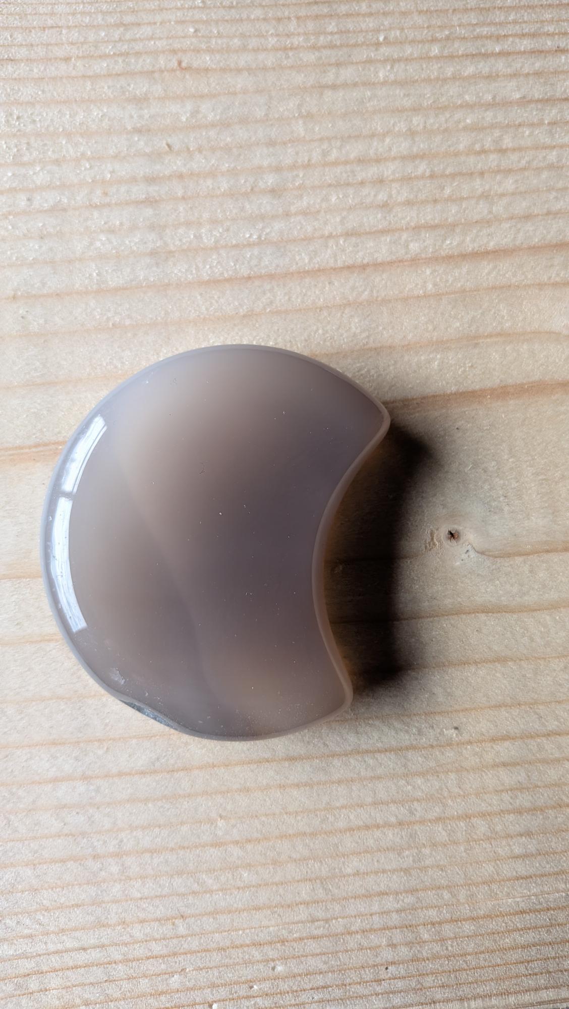 Een massief gepolijste grijze Agaat edelsteen in de vorm van een halve maan, 5 centimeter groot, gefotografeerd op een lichte houten ondergrond.