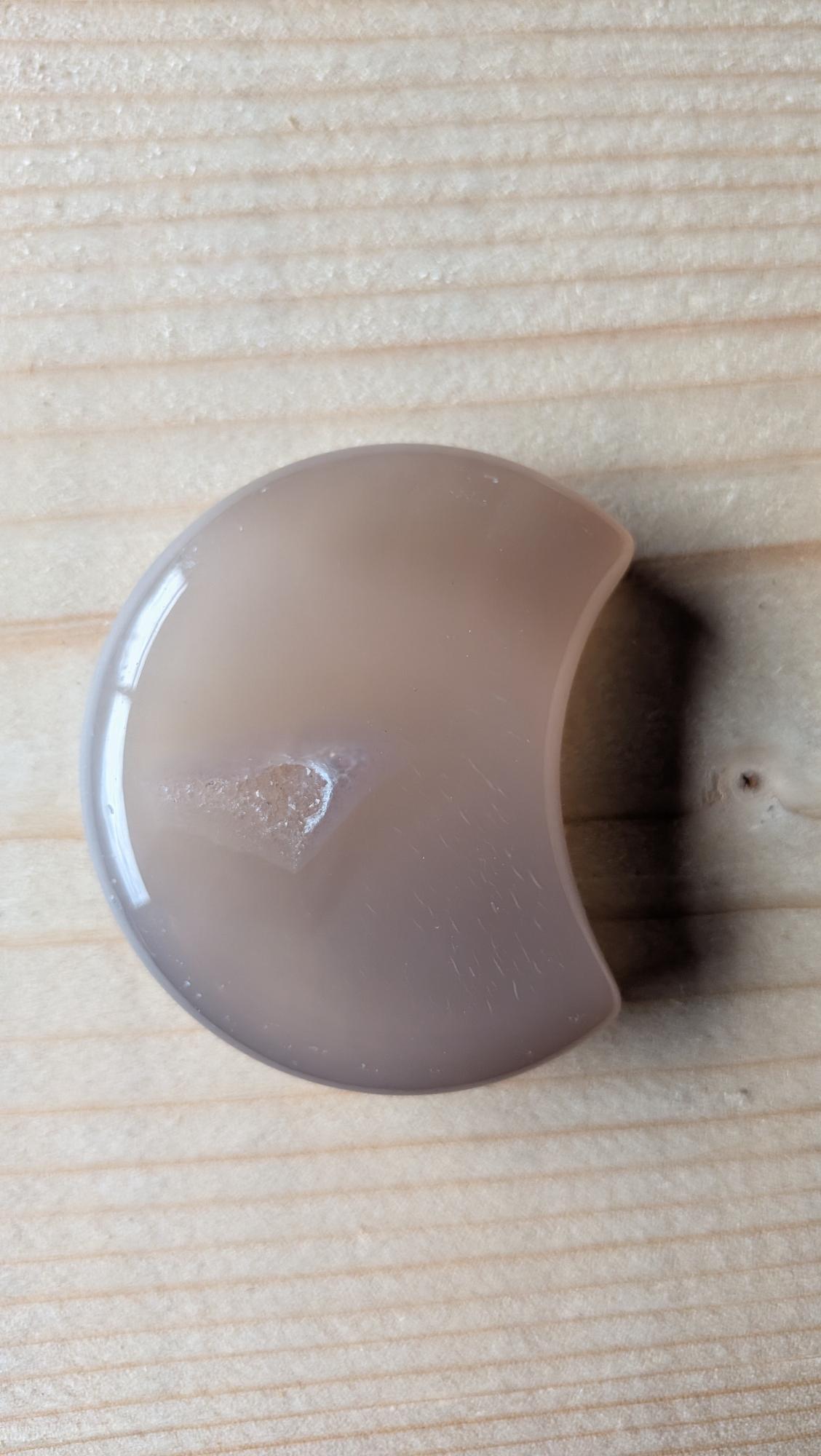 Handgeslepen grijze Agaat edelsteen in maanvorm met een kleine zichtbare kristalgeode aan de voorzijde, 5 centimeter groot.