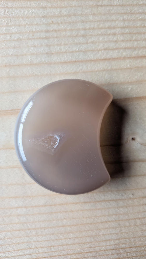 Handgeslepen grijze Agaat edelsteen in maanvorm met een kleine zichtbare kristalgeode aan de voorzijde, 5 centimeter groot.