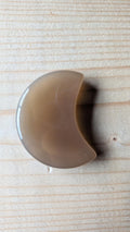 Een gepolijste grijze Agaat edelsteen in de vorm van een halve maan, 5 centimeter groot, gefotografeerd op een lichte houten ondergrond.