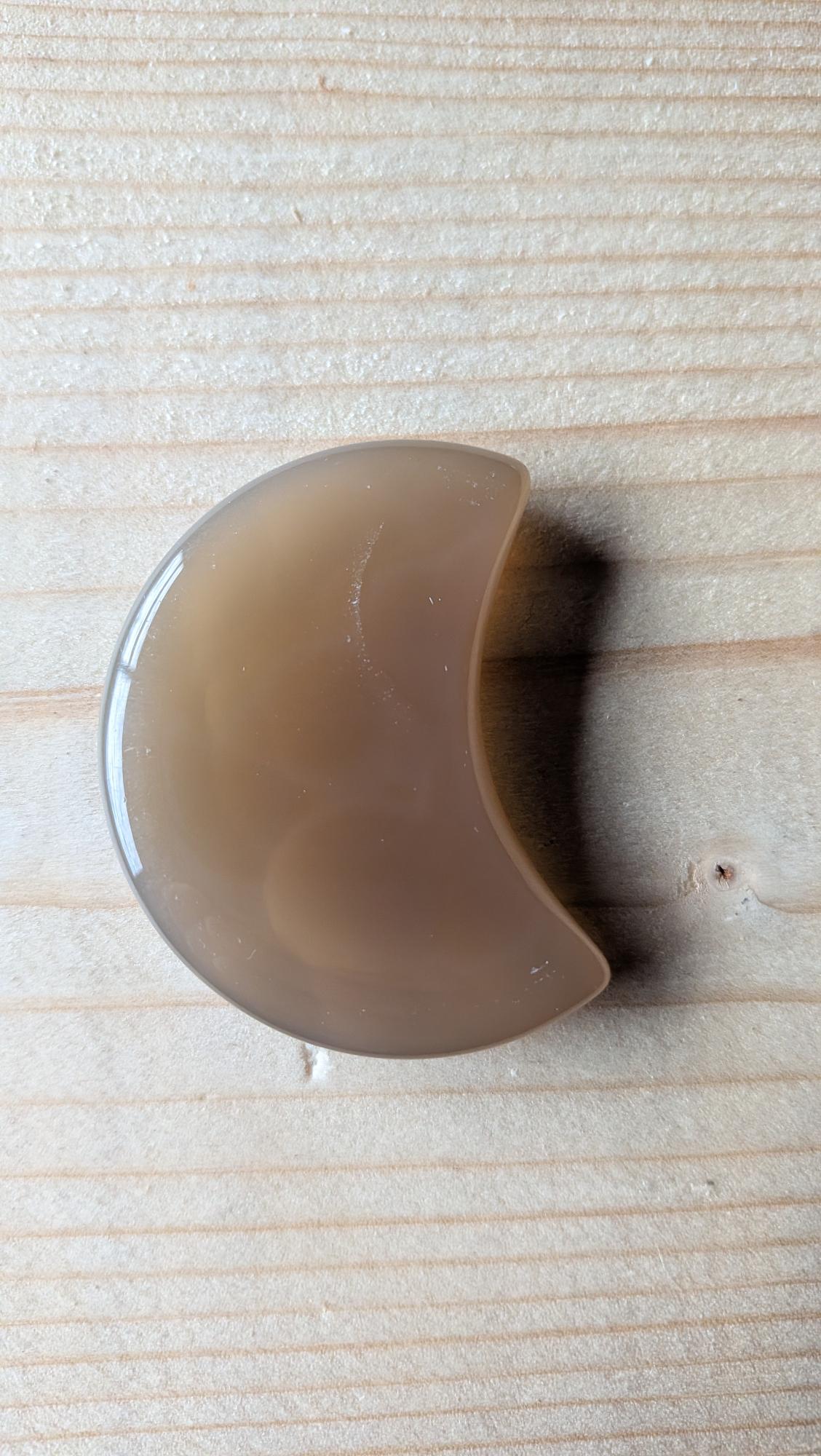 Een gepolijste grijze Agaat edelsteen in de vorm van een halve maan, 5 centimeter groot, gefotografeerd op een lichte houten ondergrond.