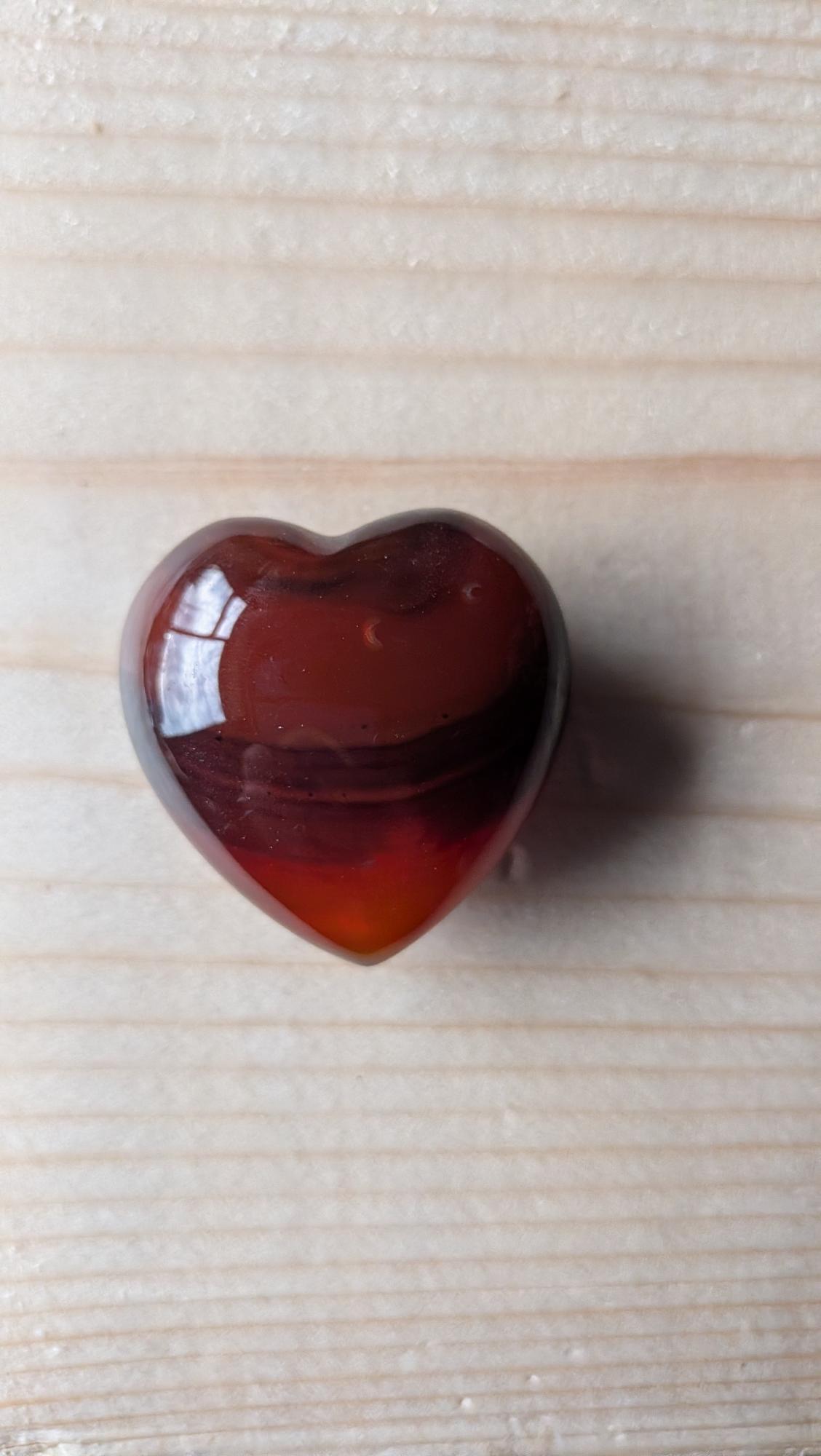 Een gepolijst edelsteen hart van Carneool met diep rode en bruine kleurbanden, 3,5 cm groot, gefotografeerd op een houten ondergrond.