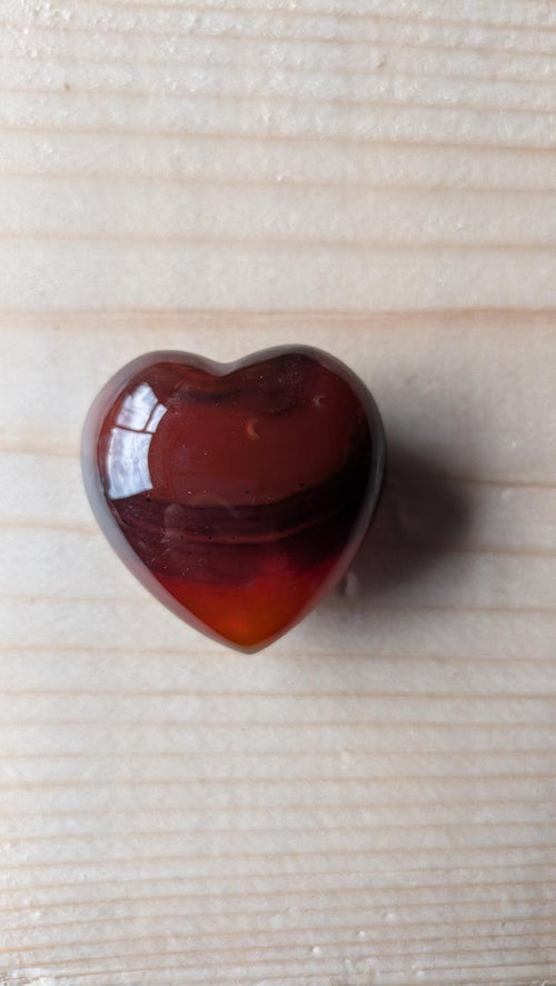 Een gepolijst edelsteen hart van Carneool met diep rode en bruine kleurbanden, 3,5 cm groot, gefotografeerd op een houten ondergrond.