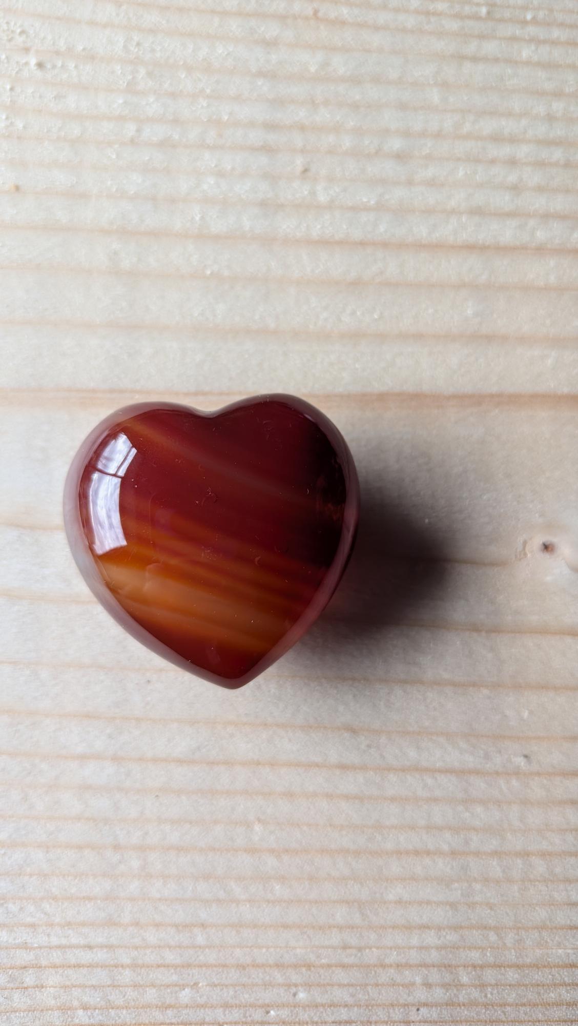 Een gepolijst Carneool hart met prachtige oranje en rode kleurbanden, 3,5 cm groot, liggend op een houten achtergrond.
