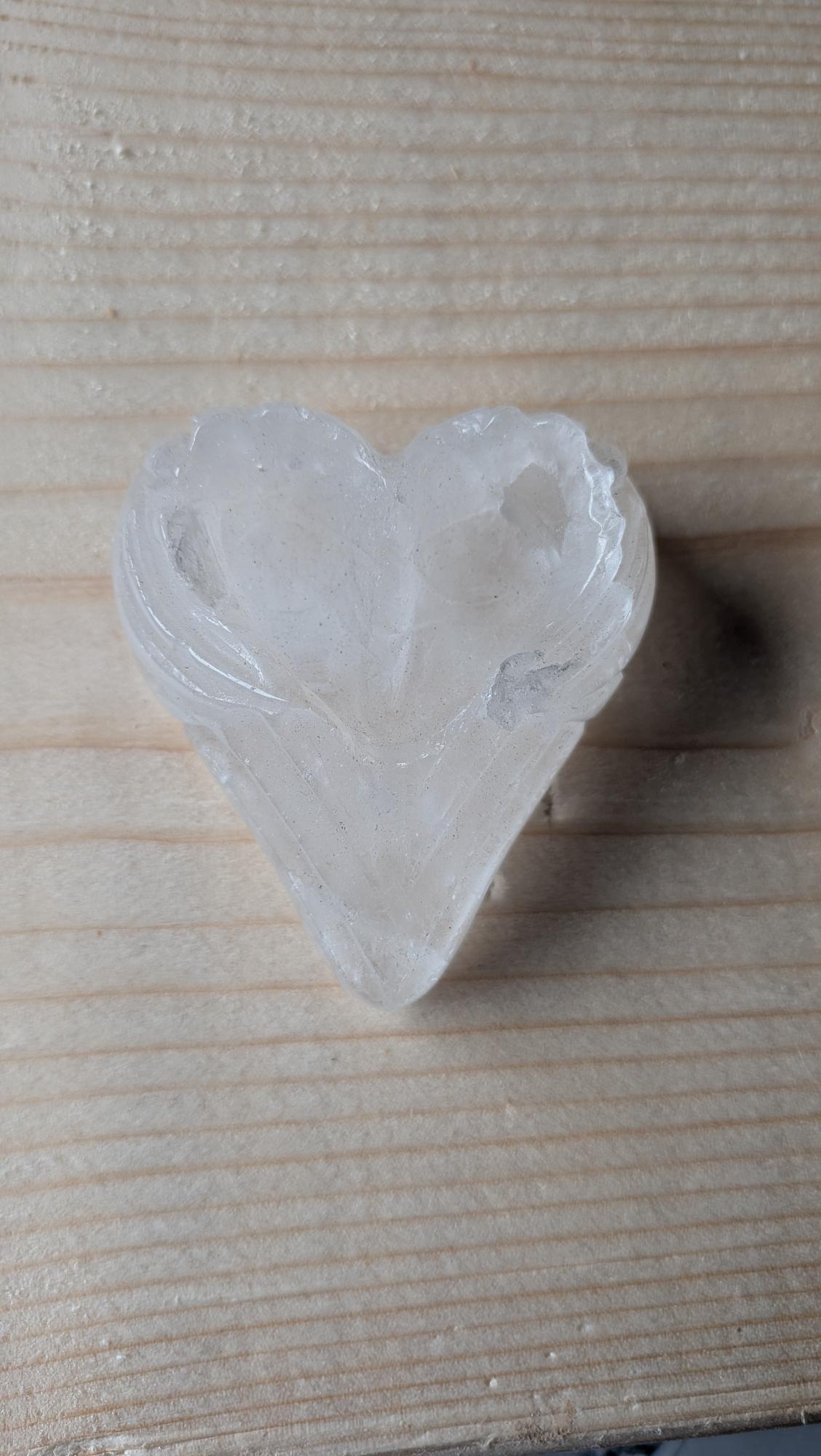 Een transparant Bergkristal hart met gedetailleerd geslepen engelenvleugels aan de bovenzijde, 6 centimeter groot, op een houten ondergrond.