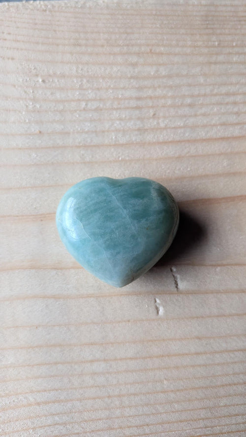 Een gepolijst groenblauw Amazoniet hartje met witachtige aders, 3,5 cm groot, gefotografeerd op een lichte houten ondergrond.