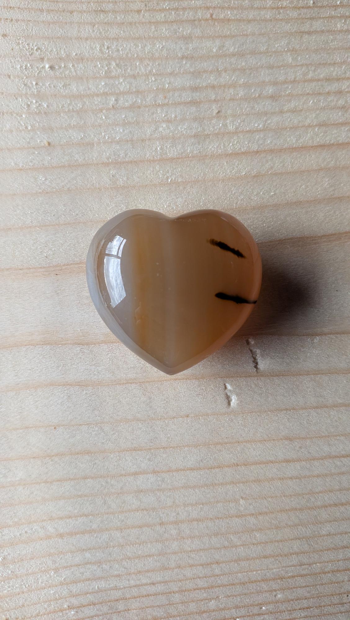Een gepolijst licht oranje/amberkleurig Carneool hartje met zwarte minerale streepjes, 3,5 cm groot, op een houten achtergrond.