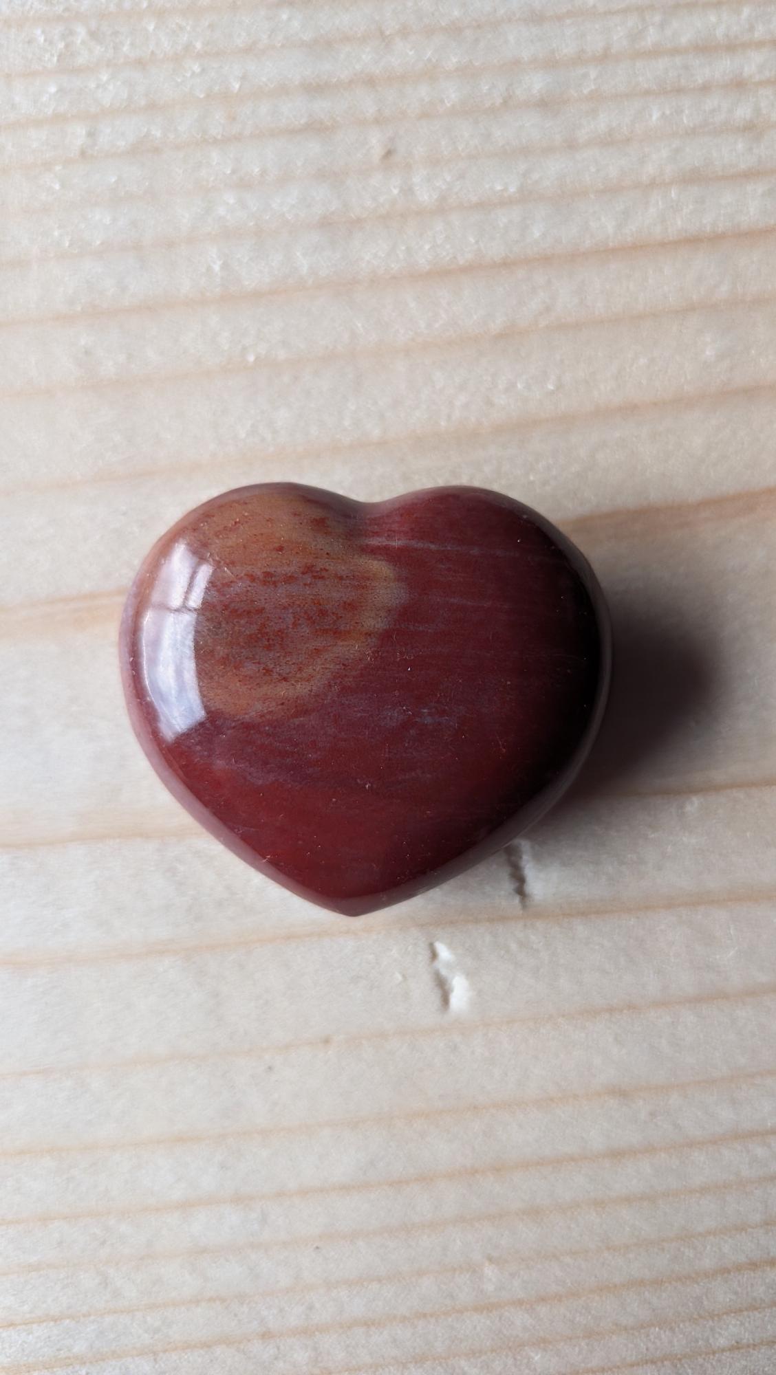 Een gepolijst dieprood edelsteen hart van Rode Jaspis met een kleine okerkleurige tekening, 3,5 cm groot, op een houten ondergrond.