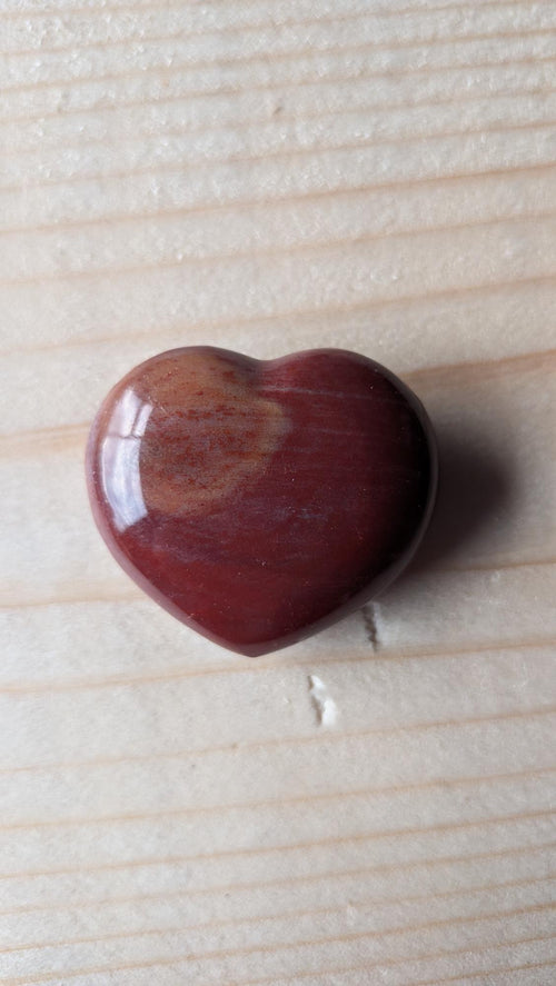 Een gepolijst dieprood edelsteen hart van Rode Jaspis met een kleine okerkleurige tekening, 3,5 cm groot, op een houten ondergrond.