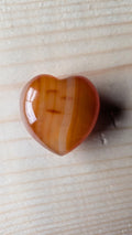 Een gepolijst, helder oranje Carneool hartje met verticale witte en amberkleurige banen, 3,5 cm groot, gefotografeerd op een houten achtergrond.