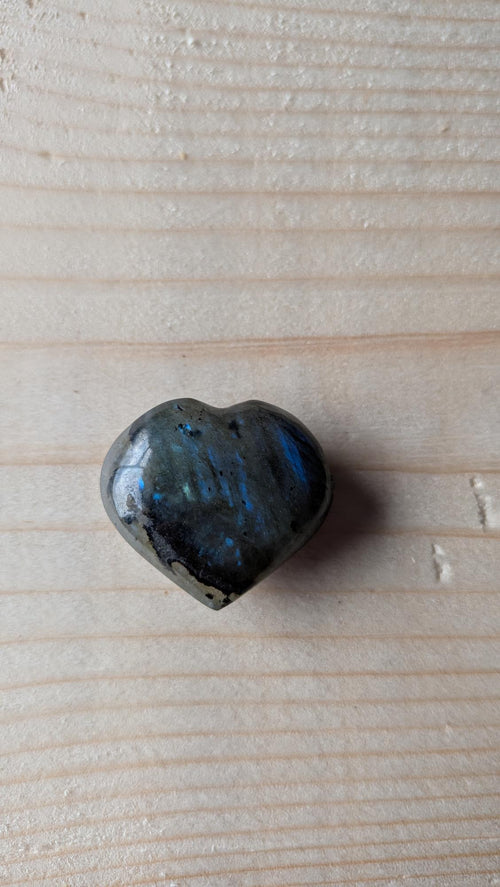 Een gepolijst donkergrijs Labradoriet hartje met een opvallende blauwe glans (labradorescentie), 3 cm groot, gefotografeerd op een lichte houten ondergrond.