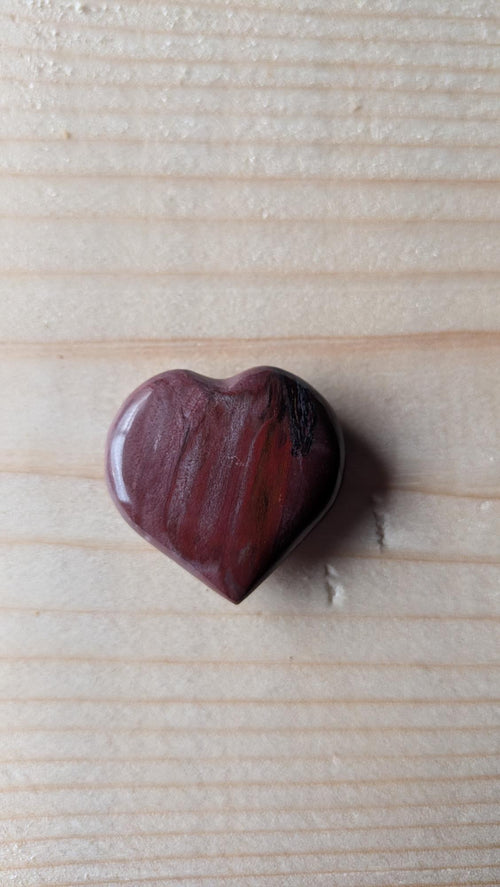 Een gepolijst hartje van Versteend Hout in diepe roodbruine tinten met zichtbare houtstructuur, 3 cm groot, gefotografeerd op een houten ondergrond.