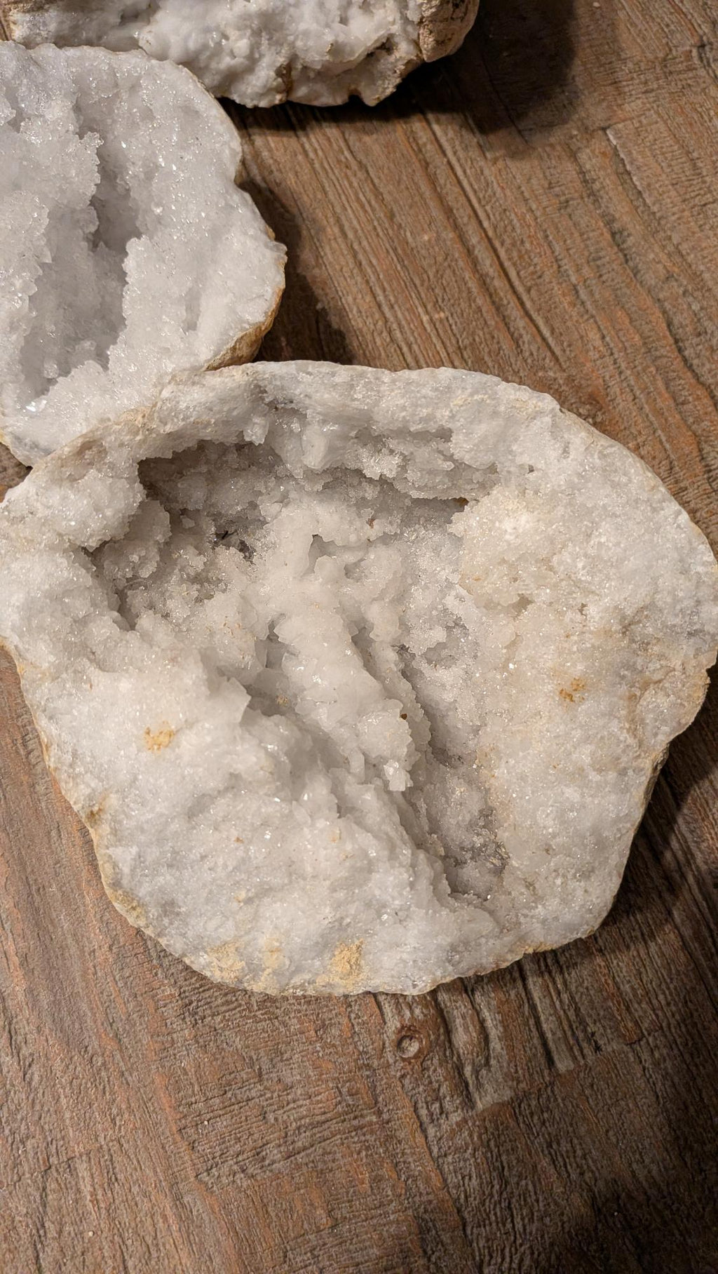 Brede, platte witte kristalgeode van 1155 gram met een groot oppervlak aan schitterende kristallen, liggend op een houten ondergrond.