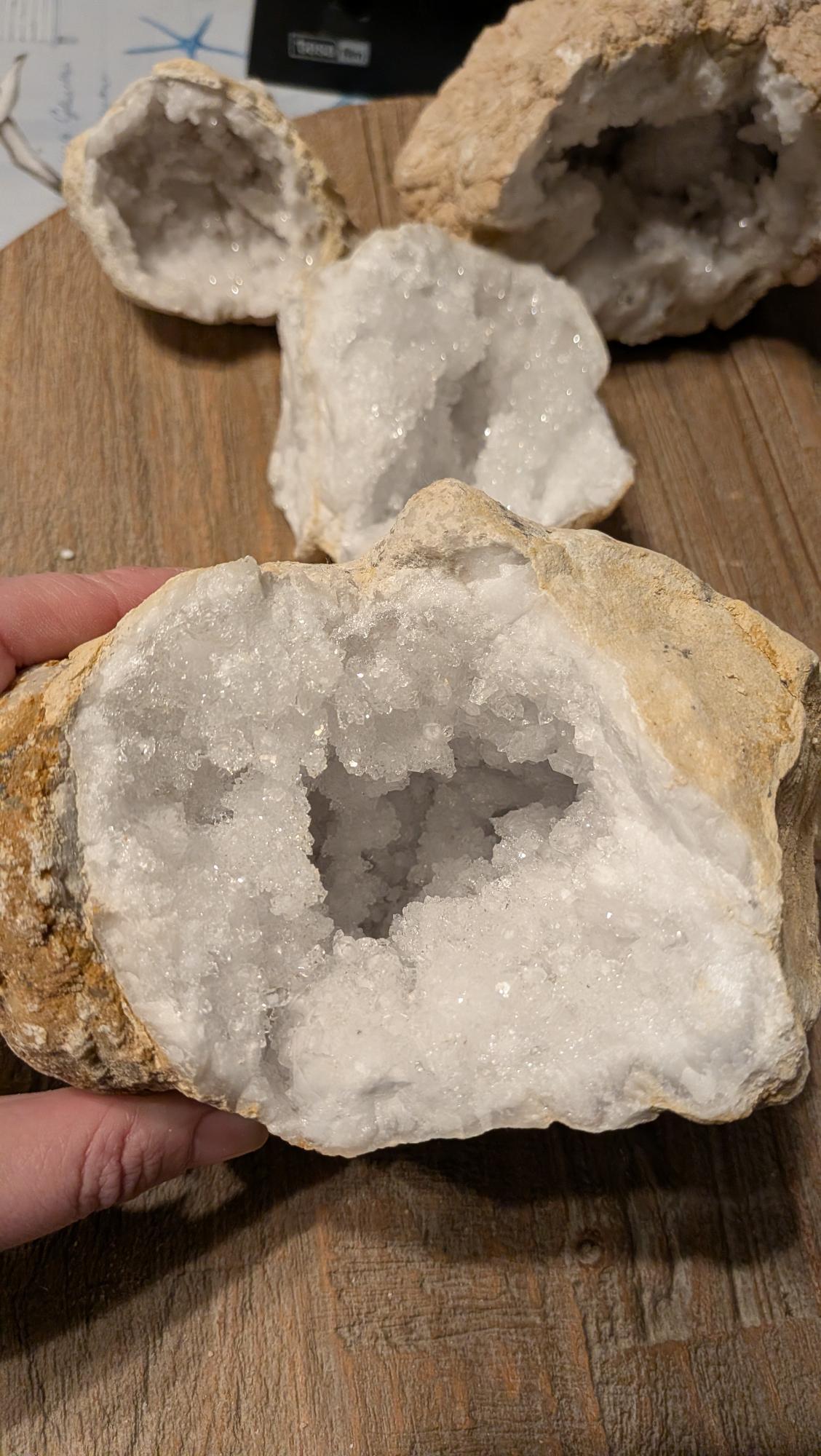 Zeer grote, zware kristalgeode van 1677 gram met een diepe binnenzijde vol witte kristallen, liggend op een houten tafel.