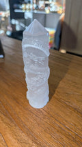 Witte satijnachtige Seleniet kristal toren van 11 cm met een gedetailleerde inkerving van een draak op een houten ondergrond.