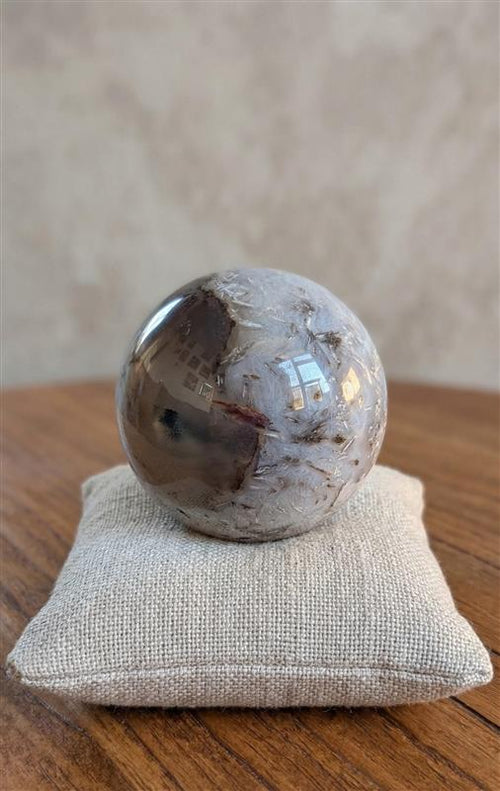 Gepolijste Agaat Geode bol van 6,5 cm met een grote, sprankelende quartz kristallen holte (druse) op een beige kussentje, gewicht 369 gram.