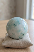 Lichtblauwe Phoenix Stone bol van 7 cm met turquoise spikkels op een linnen kussentje, gewicht 477 gram. Kristal voor balans en wedergeboorte.