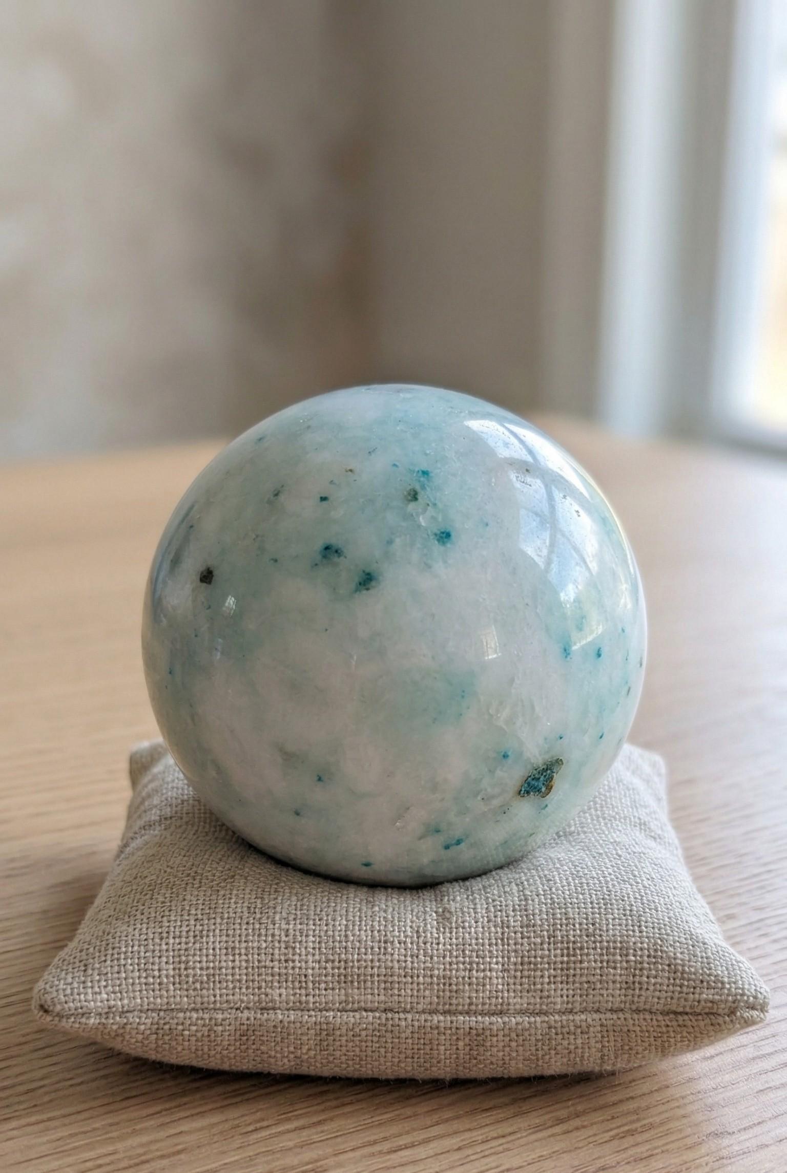 Lichtblauwe Phoenix Stone bol van 7 cm met turquoise spikkels op een linnen kussentje, gewicht 477 gram. Kristal voor balans en wedergeboorte.
