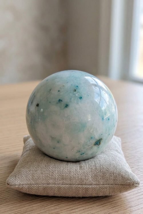 Lichtblauwe Phoenix Stone bol van 7 cm met turquoise spikkels op een linnen kussentje, gewicht 477 gram. Kristal voor balans en wedergeboorte.