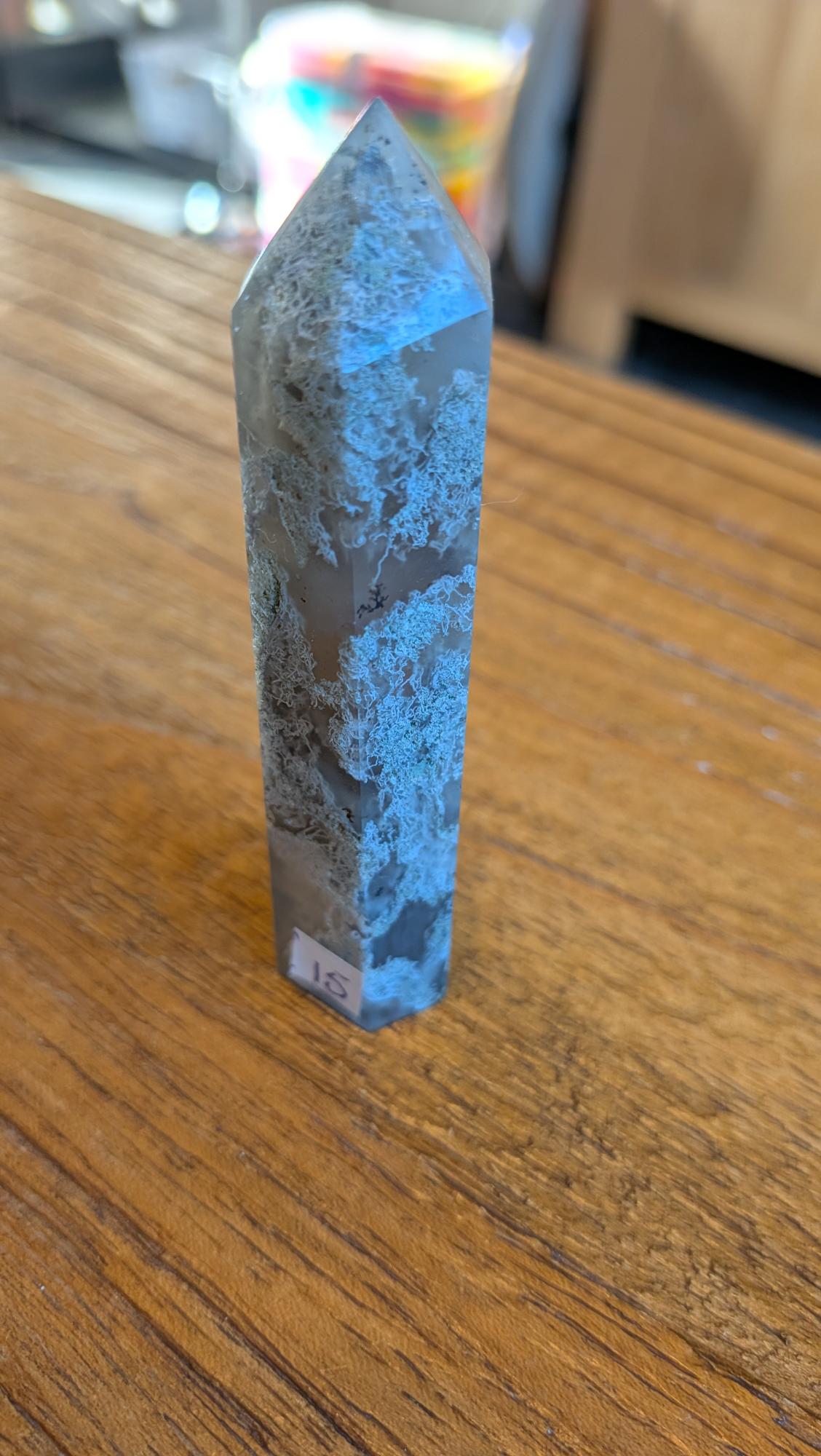 Slanke Mos Agaat kristal toren van 9 cm met witte en diepgroene mos-achtige patronen op een houten ondergrond.