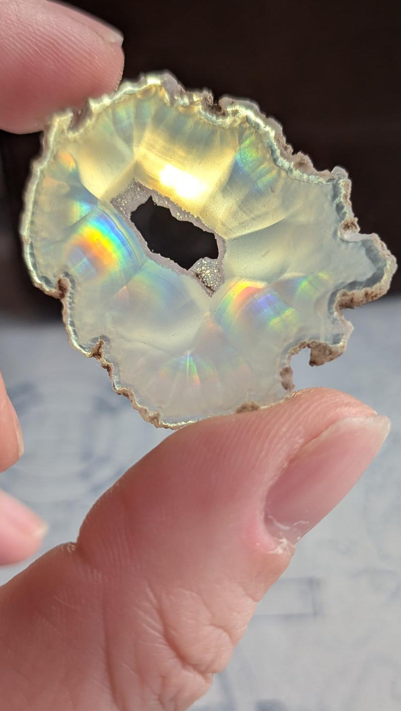 Compact plakje heldere Iris Agaat uit Java (4 cm) met een scherpe regenboogflits en transparante kristalstructuur, tegen het licht gehouden.