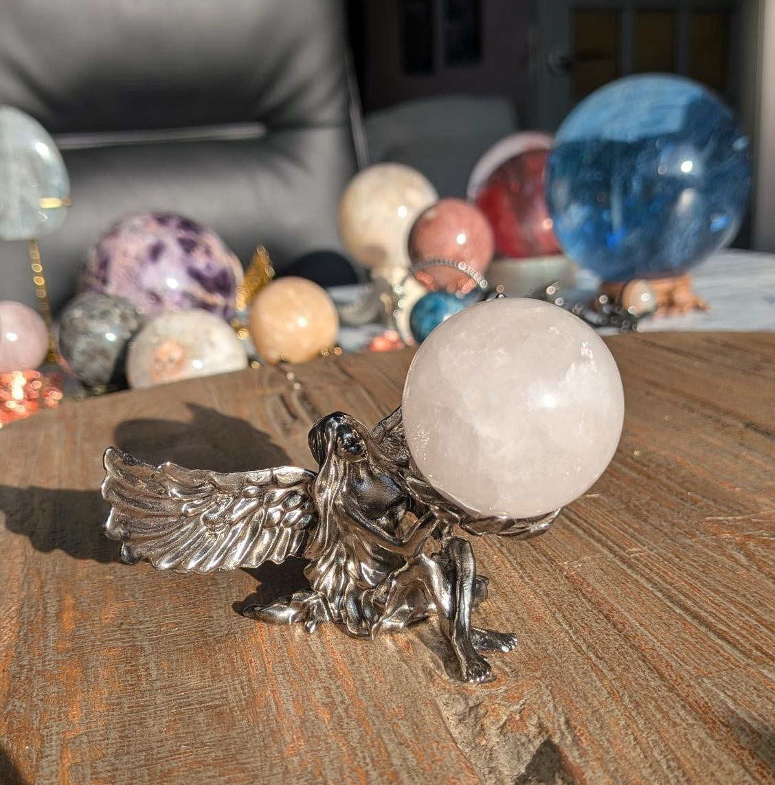 Een lichtroze Rozenkwarts bol van 4 cm die wordt vastgehouden door een gedetailleerde zilverkleurige engel-houder op een rustieke houten tafel.