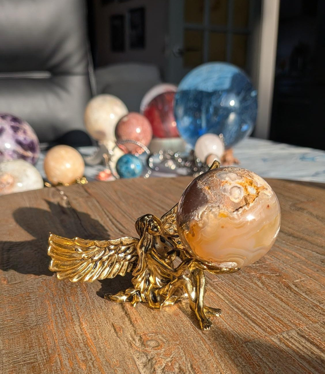 Een Bloemagaat bol met beige en bruine bloempatronen, gedragen door een gedetailleerde goudkleurige engel-houder op een rustieke houten ondergrond.