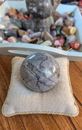 Gepolijste grijze Picasso Jaspis bol van 4,5 cm met abstracte zwarte lijnen op een beige kussentje, prijs 16,50 euro. Houders apart verkrijgbaar.