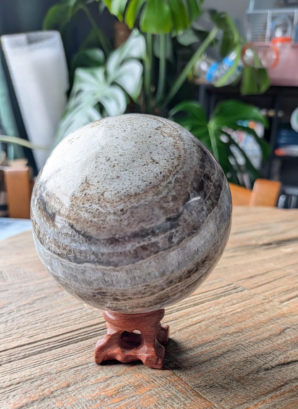 Een grote, rond geslepen Marokkaanse jaspis bol met natuurlijke grijze en bruine banden, rustend op een kleine houten standaard op een houten tafel.