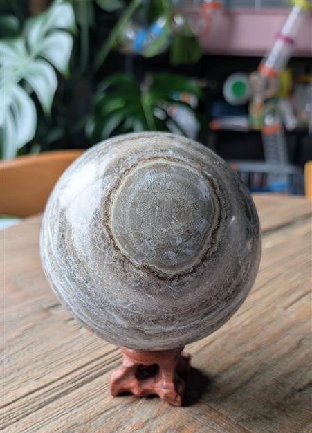 Een grote, rond geslepen Marokkaanse jaspis bol met natuurlijke grijze en bruine banden, rustend op een kleine houten standaard op een houten tafel.