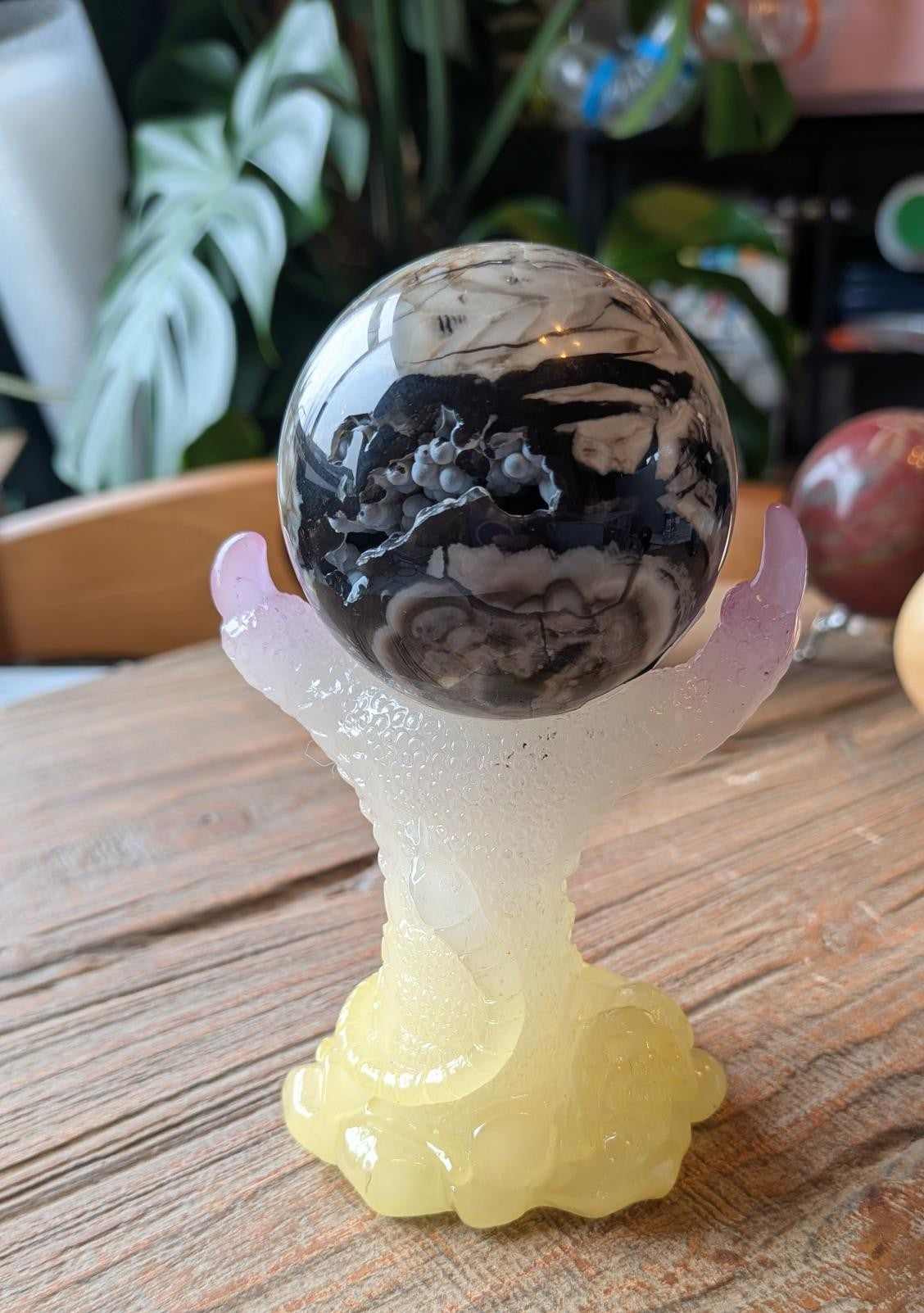 Een donkere Vulkaan Agaat bol op een kleurrijke geel-paarse metalen houder in de vorm van een klauw met een slang, gefotografeerd op een houten ondergrond.