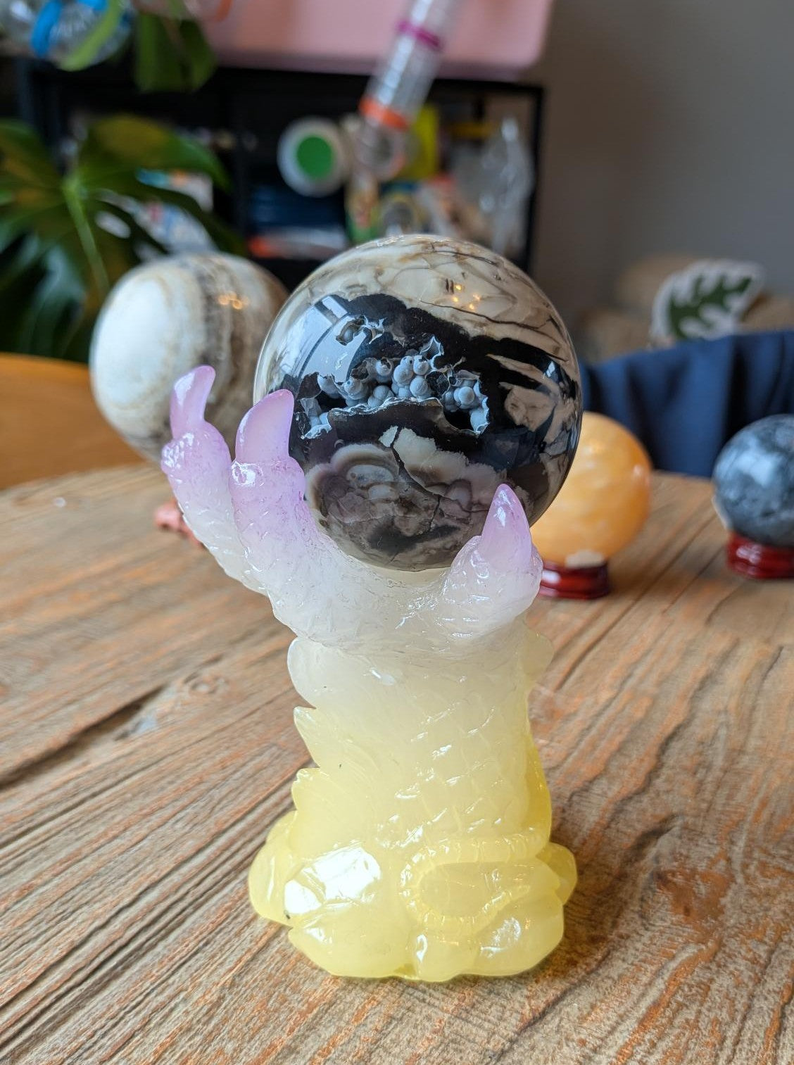 Een donkere Vulkaan Agaat bol op een kleurrijke geel-paarse metalen houder in de vorm van een klauw met een slang, gefotografeerd op een houten ondergrond.