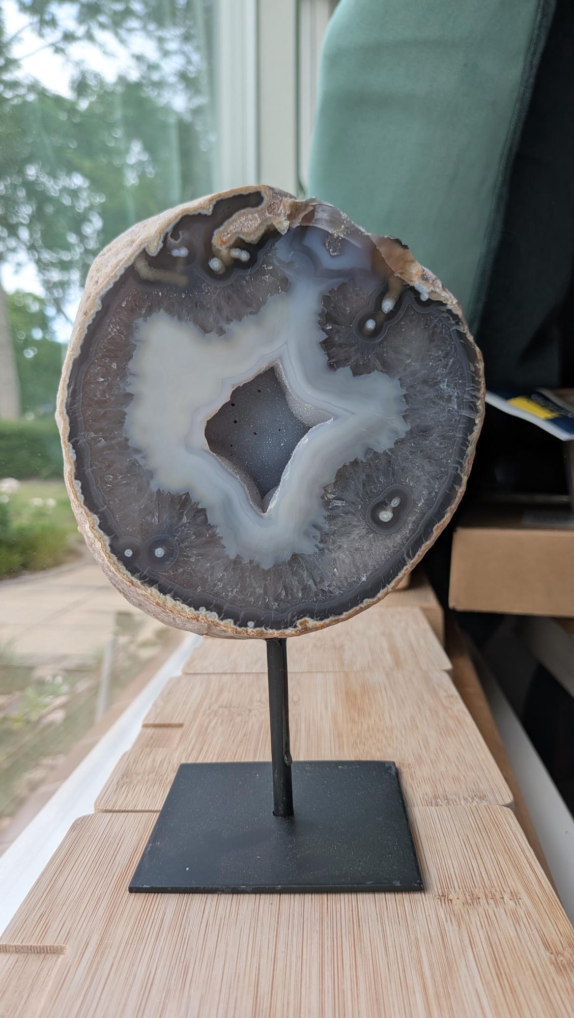 Een hand houdt een grote Agaat-geode van 1,7 kg op een zwarte standaard vast, met een opvallend ster-vormig kristalhart in het midden".