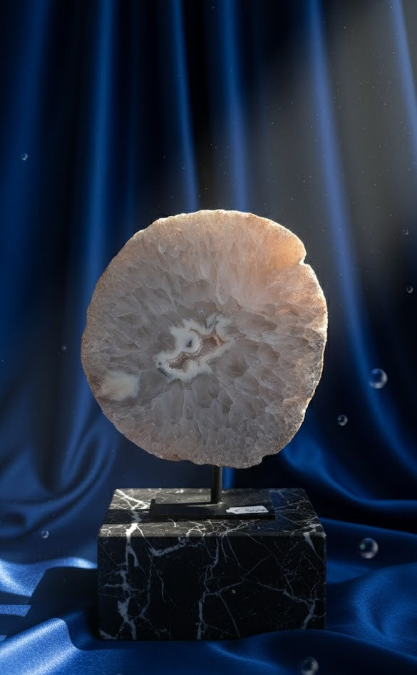 Grote, ronde Agaat schijf van 1049 gram met een wit-roze kristalcentrum op een zwarte metalen standaard, gepresenteerd op een marmeren blok tegen een diepblauwe satijnen achtergrond.