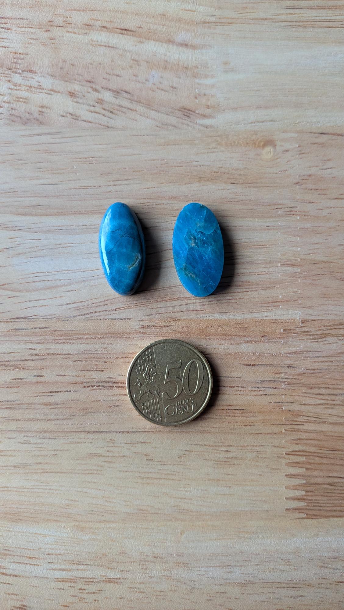 Twee ovale, gepolijste blauwe apatiet edelstenen met een levendige oceaanblauwe kleur naast een 50 eurocent munt op een houten achtergrond.