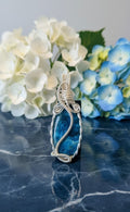 Rechtopstaande handgemaakte verzilverde wire-wrapped hanger met een ruwe blauwe apatiet edelsteen, gepresenteerd op een donkere marmeren ondergrond met witte en blauwe hortensia's op de achtergrond.