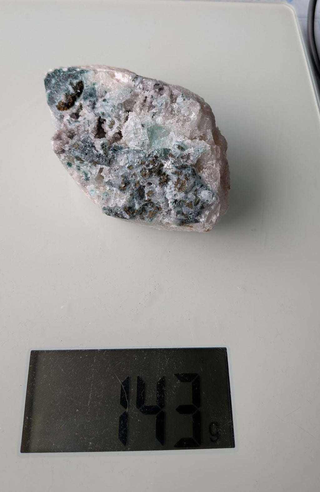 Een exclusief, ruw Apofylliet cluster uit Nasik, India, met glasachtige heldere kristallen op een zachtroze Stilbiet-matrix, gewogen op een digitale weegschaal die 143 gram aangeeft.