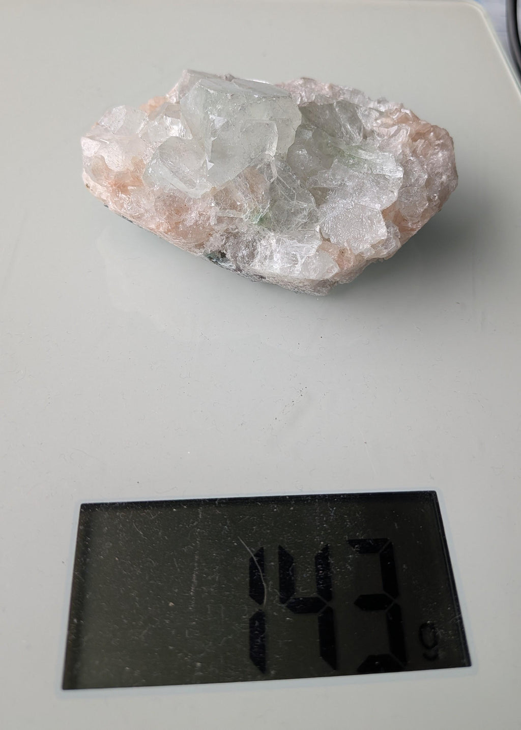 Een exclusief, ruw Apofylliet cluster uit Nasik, India, met glasachtige heldere kristallen op een zachtroze Stilbiet-matrix, gewogen op een digitale weegschaal die 143 gram aangeeft.