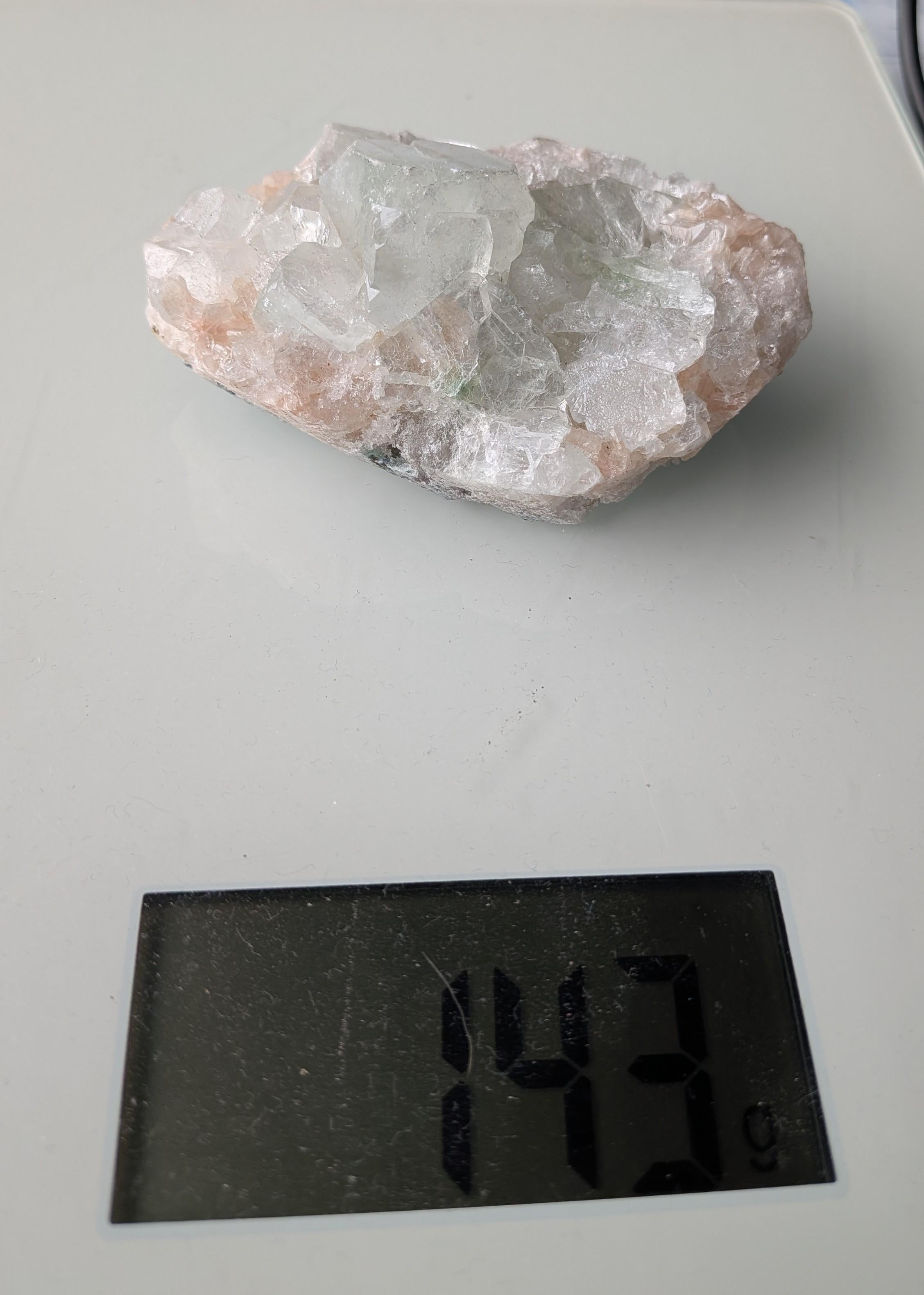 Een exclusief, ruw Apofylliet cluster uit Nasik, India, met glasachtige heldere kristallen op een zachtroze Stilbiet-matrix, gewogen op een digitale weegschaal die 143 gram aangeeft.
