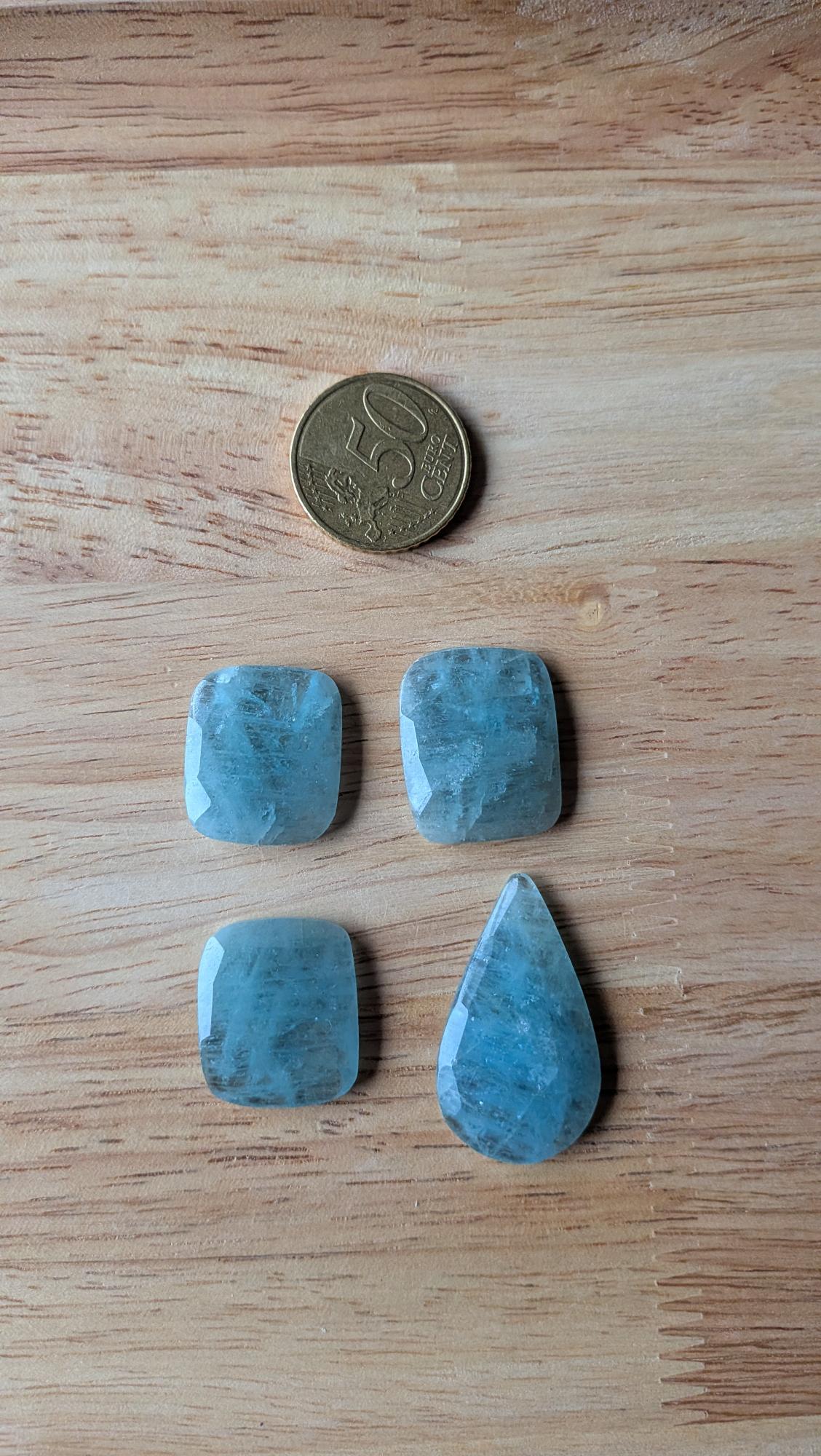 Vier facetgeslepen lichtblauwe Aquamarijn edelstenen in kussenvorm en druppelvorm op een houten ondergrond naast een 50 cent muntstuk.